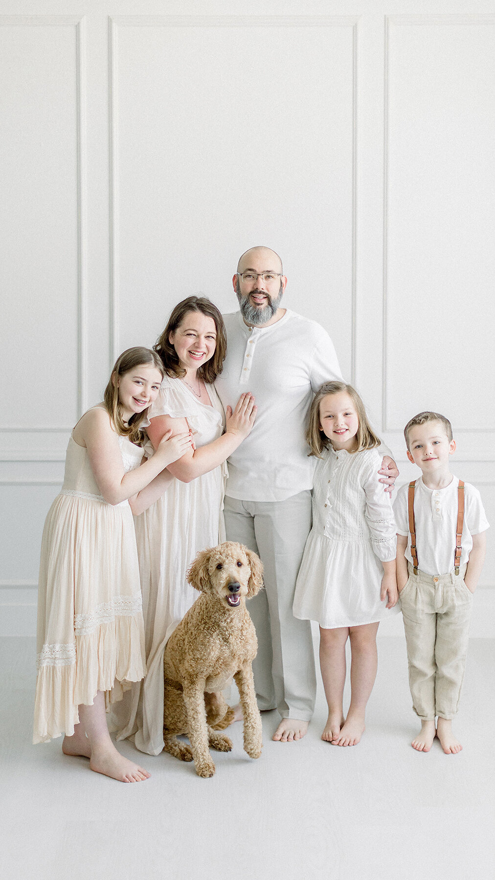 A family portrait of 5 with their dog while standing together posing by a window at a photography studio in the Fort Worth area by a luxury family photographer.