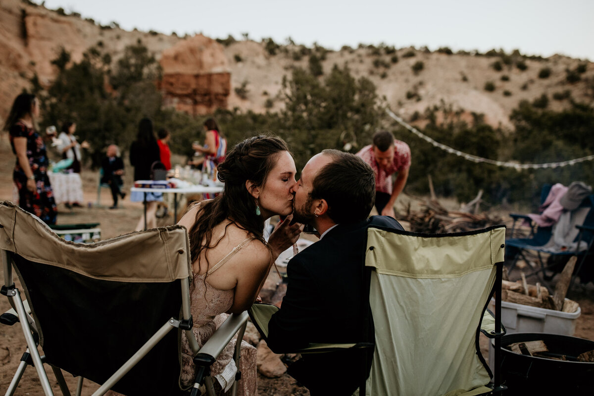 bride and groom sitting by campfire kissing