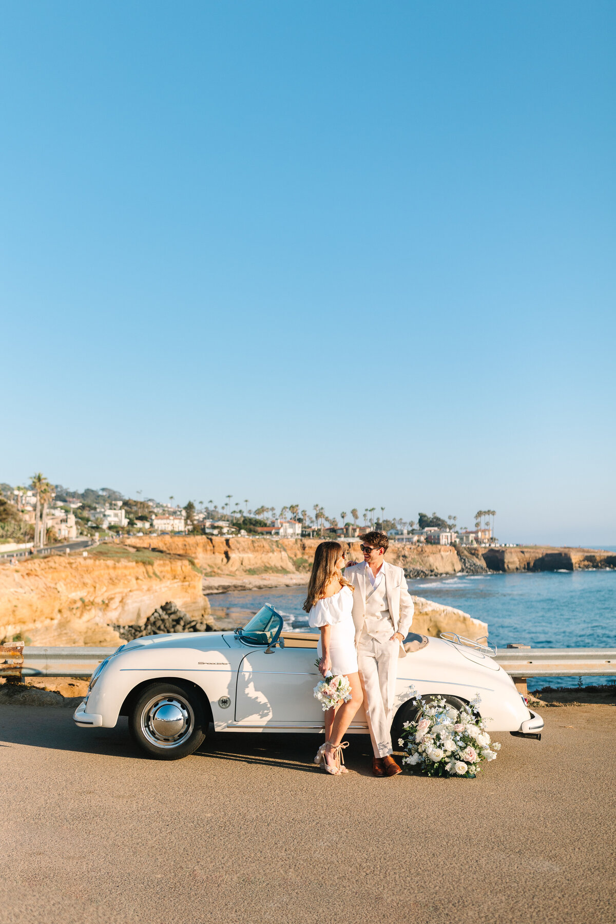 bride and groom standing in front of white vintage car by the beach.