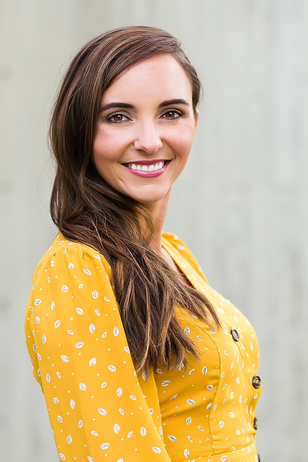Woman in thirties wearing yellow and posing for headshot in front of a cement wall