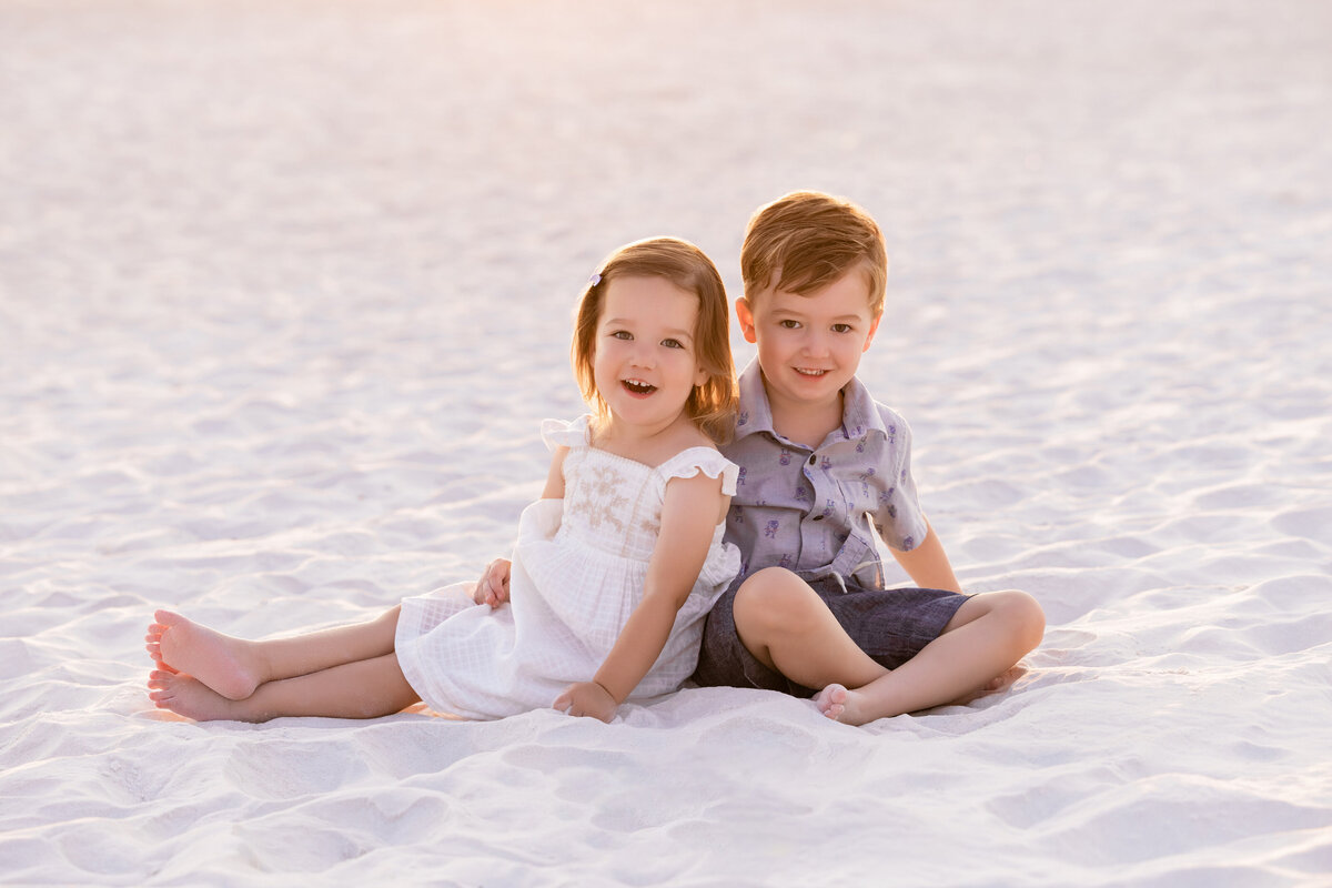 Two kids sitting in the sand together smiling