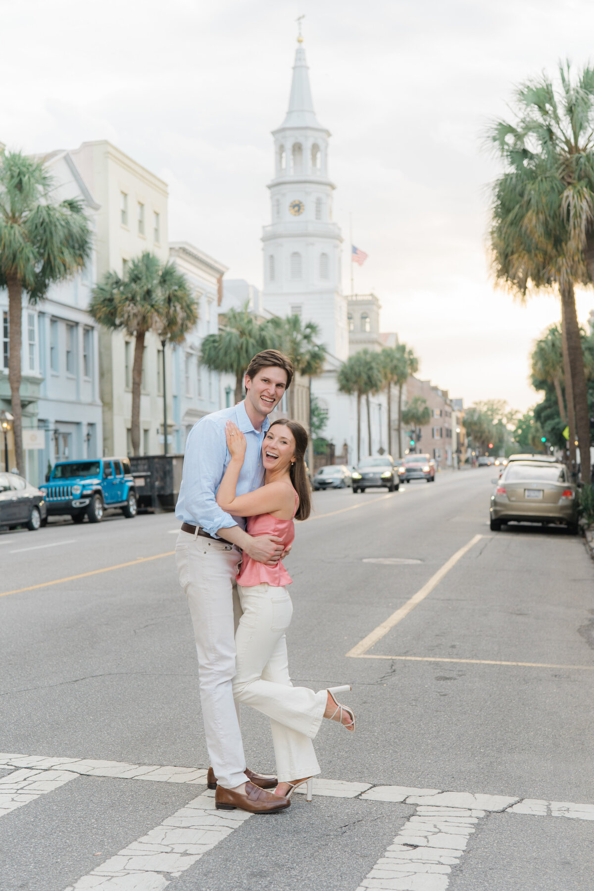 Broad street in Charleston. Palm trees and big church steeple. Couple standing in the crosswalk for cute engagement photo. Downtown Charleston engagement session. Kailee DiMeglio Photography.