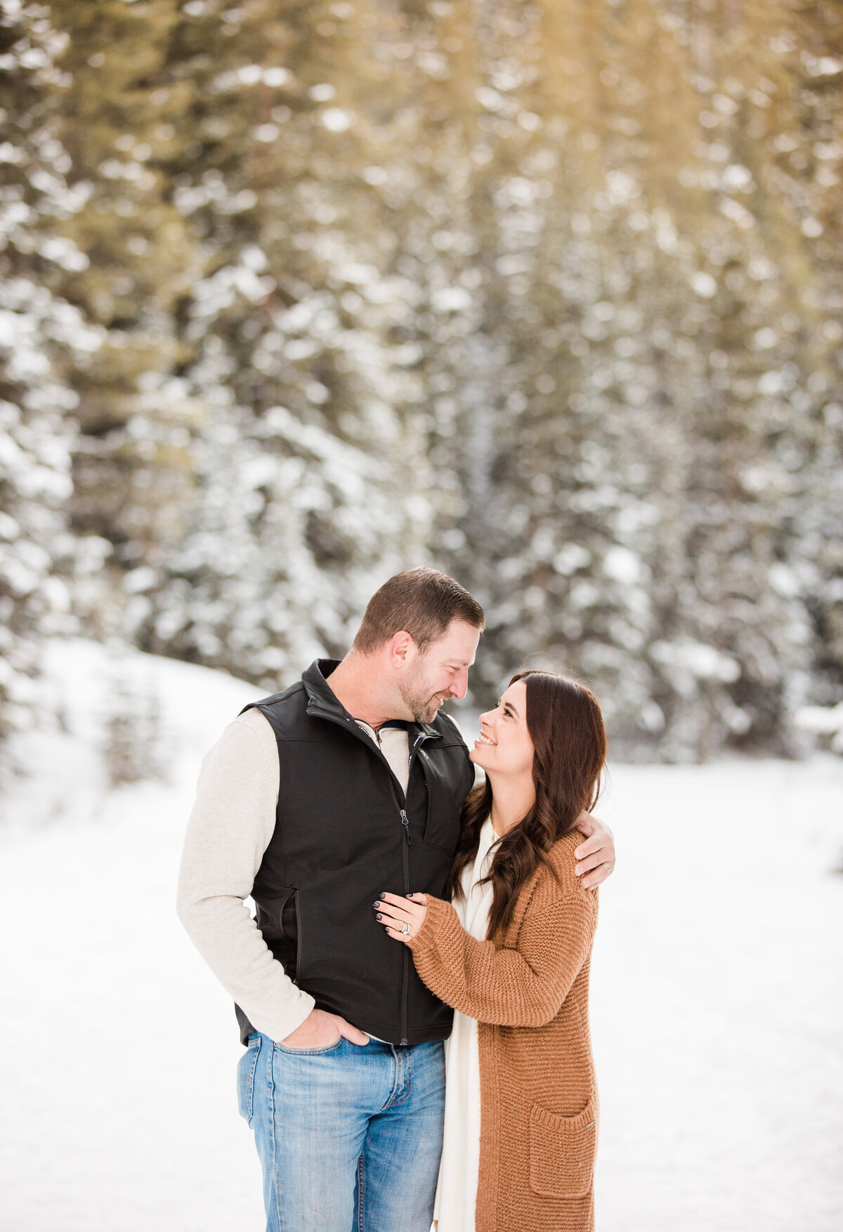 A man and woman are standing closely together looking at each other. They are in a winter scene of pine trees and snow all around