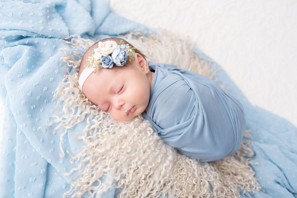 A sleeping newborn baby on a blue blanket in a blue swaddle and matching floral headband posed by an Atlanta newborn photographer