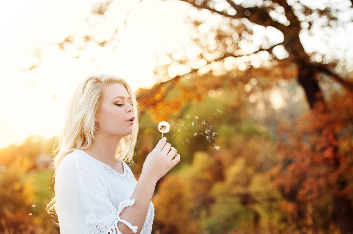girl blowing dandelion in colorful fall tress