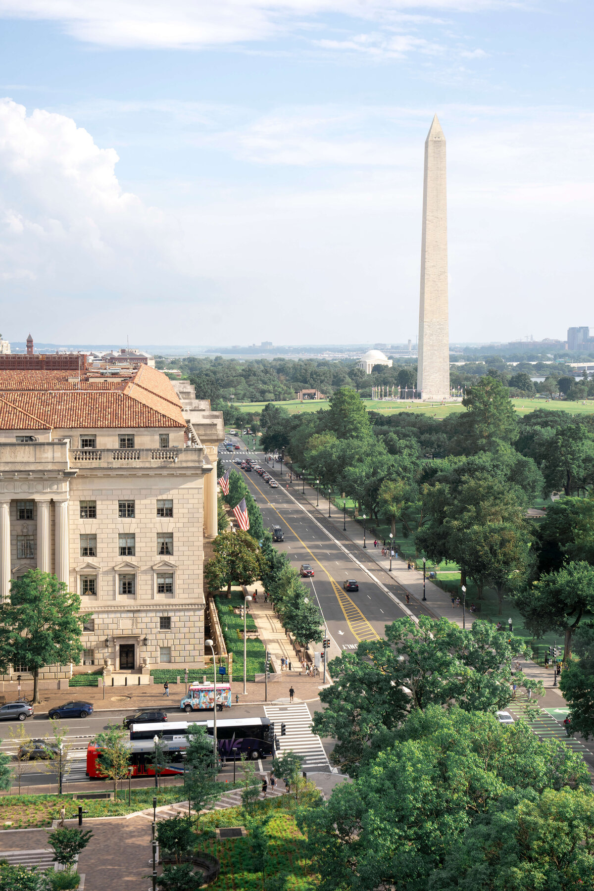 Aerial view of a city street lined with trees and surrounded by historic buildings. The Washington Monument stands prominently in the background under a partly cloudy sky. Buses and cars travel along the road.