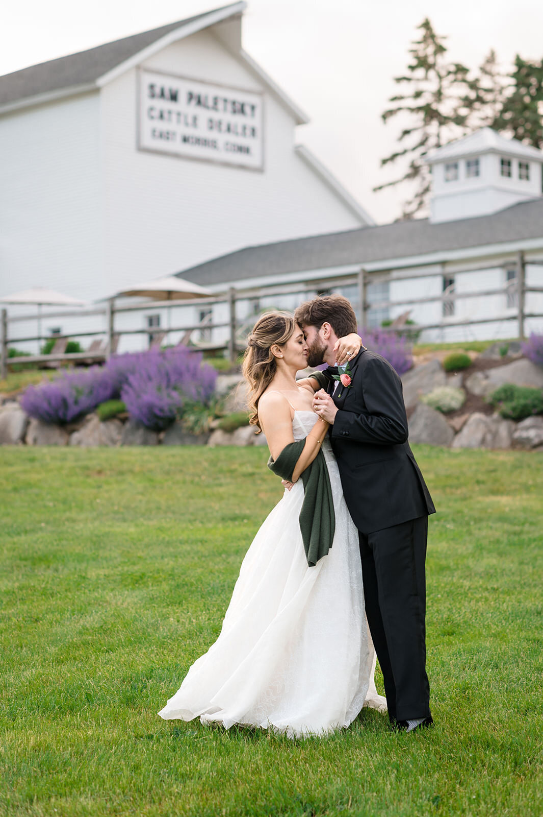A bride and groom sharing a kiss on a lawn in front of a building with a sign that reads "Sam Paley, Cattle Dealer."