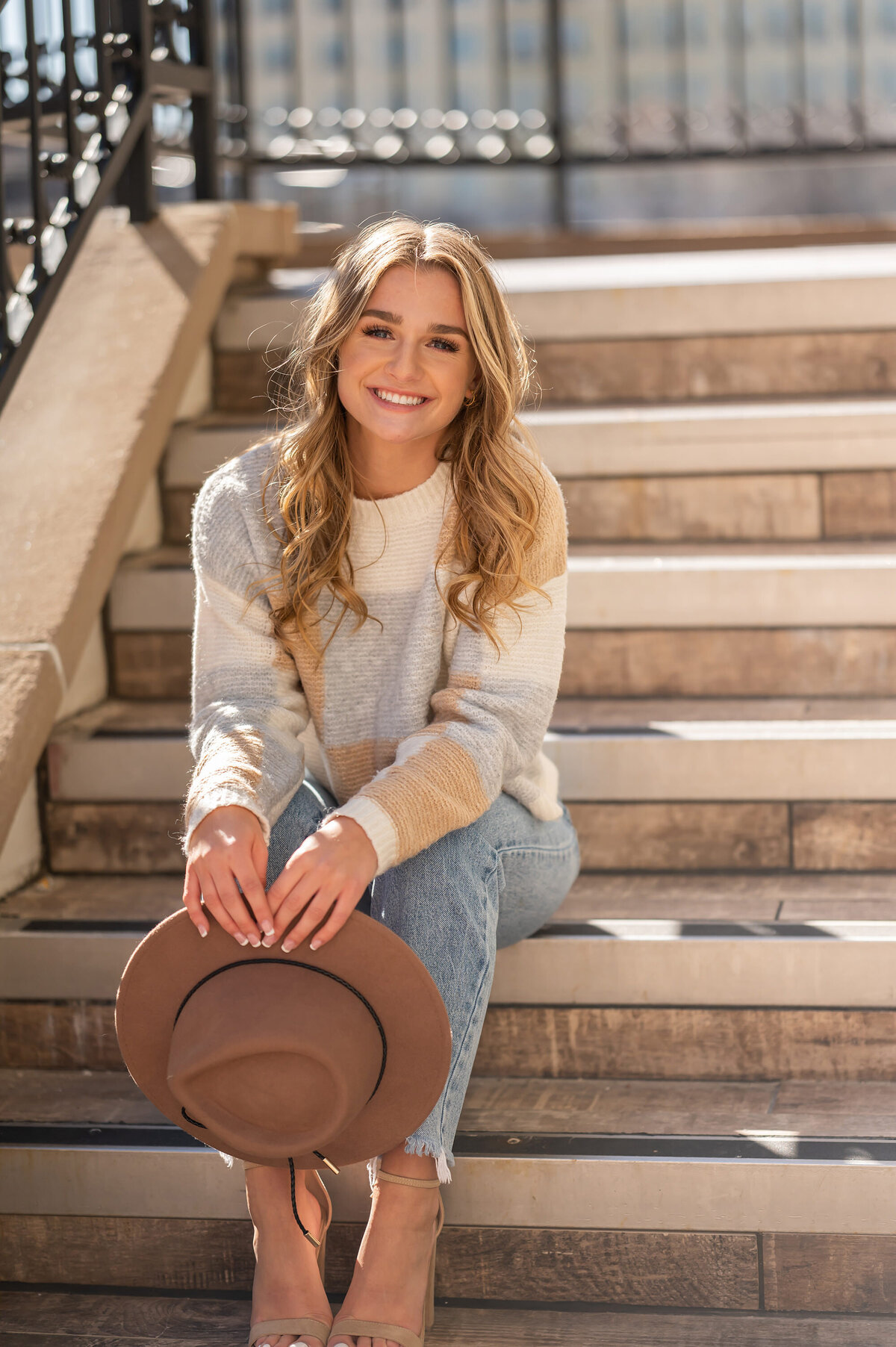 A young woman from New Berlin West High School sits at the base of a stone staircase in Downtown Waukesha wearing jeans and  sweater while holder her hat.