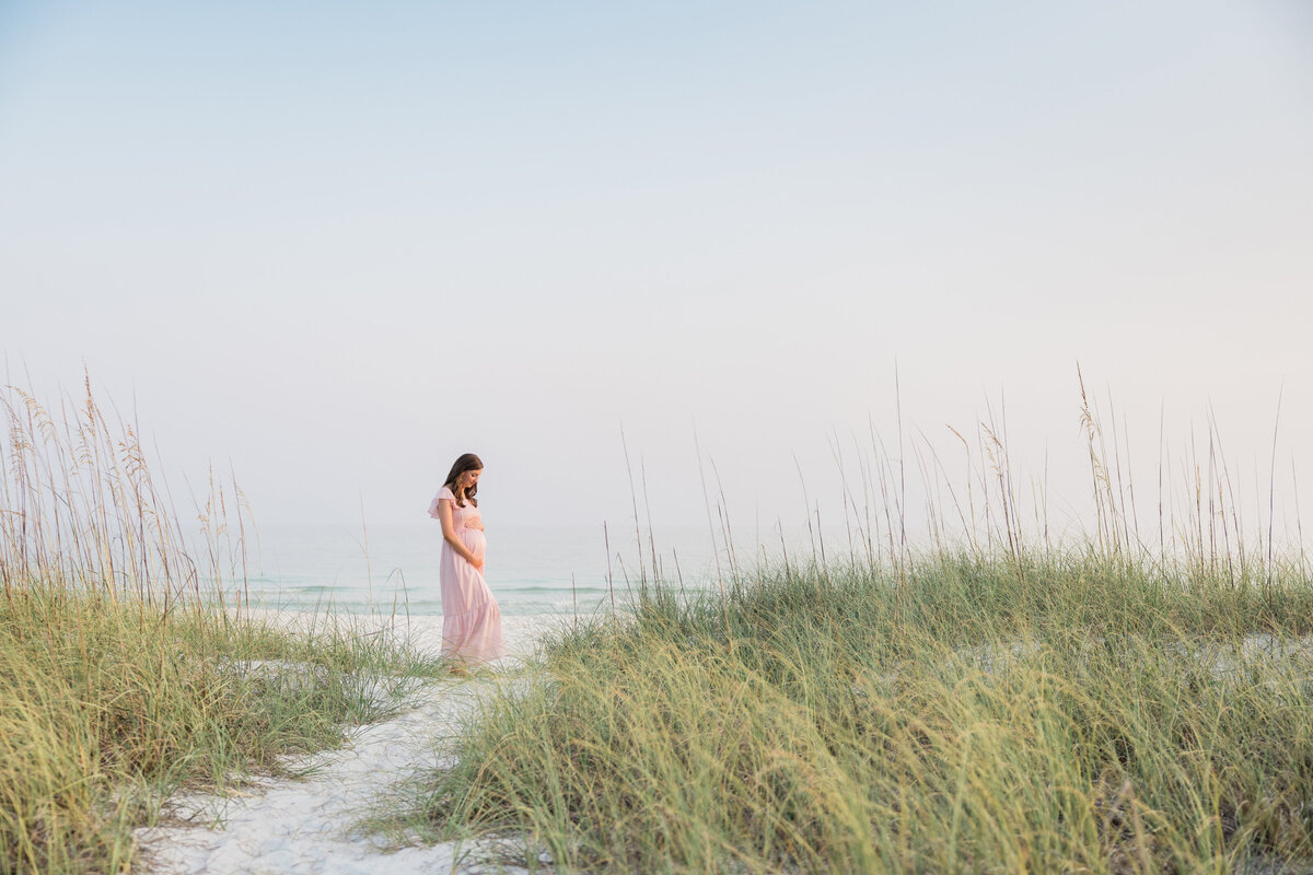 A maternity portrait of a woman standing near a sand dune in Inlet Beach Florida, wearing a pink dress.