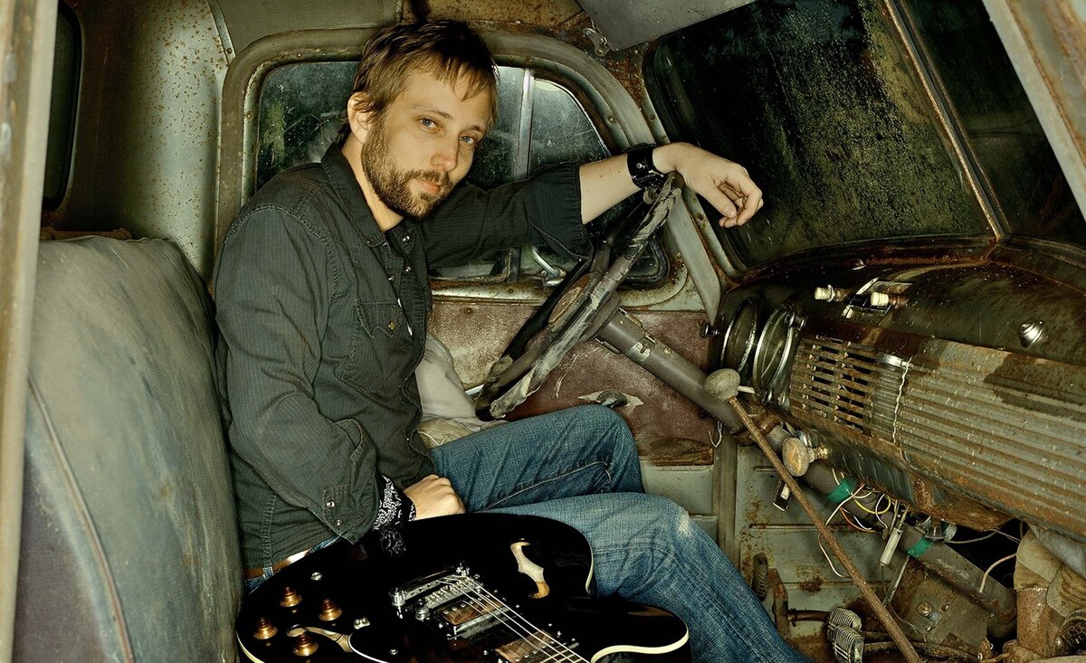 Male musician portrait Ryan McMahon wearing green dress shirt blue jeans sitting inside old truck cab with black electric guitar