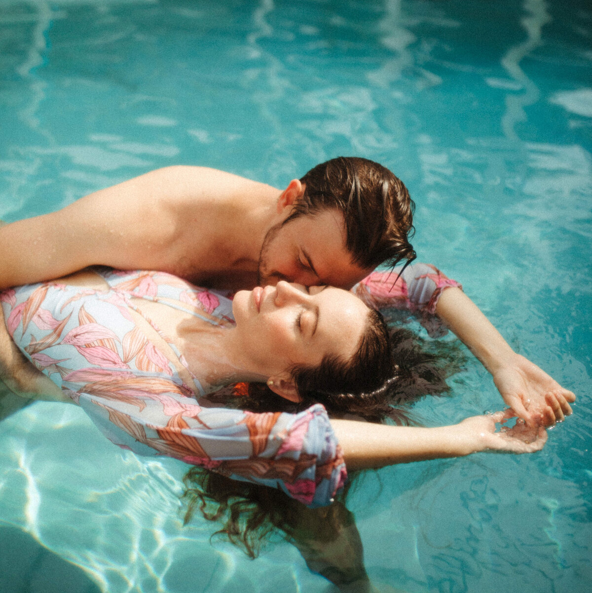 A girl in a vintage swimsuit is floating in a swimming pool while a guy is holding her and kissing her.