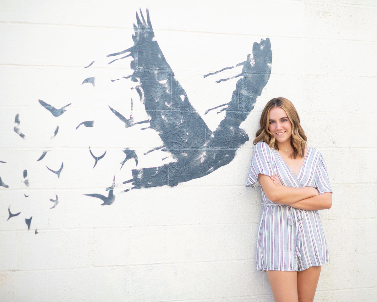 High school senior girl smiles for photo in front of white wall with bird painting