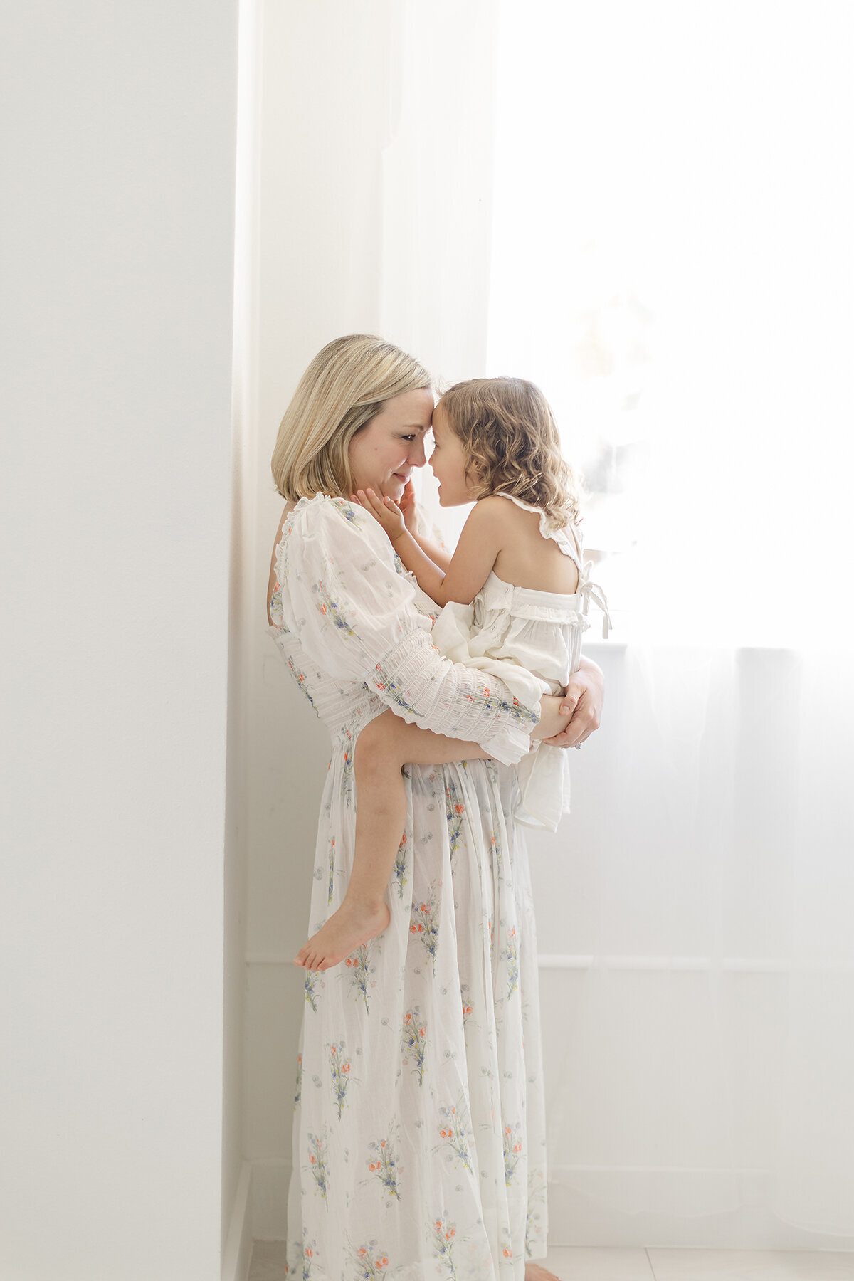 A mother standing by a window in a photography studio as she holds her toddler girl close.