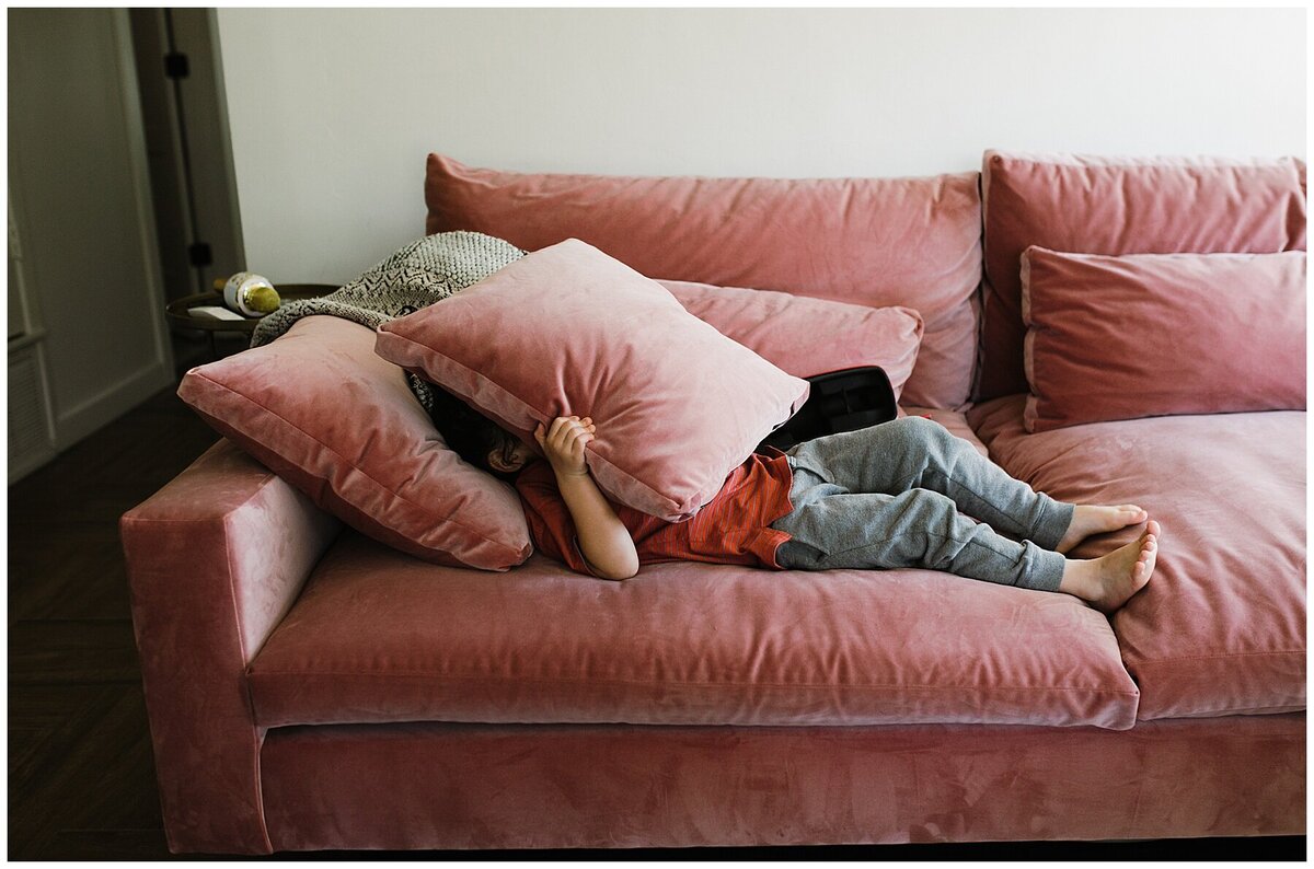 Child hiding under a pink pillow lying on a velvet pink sofa