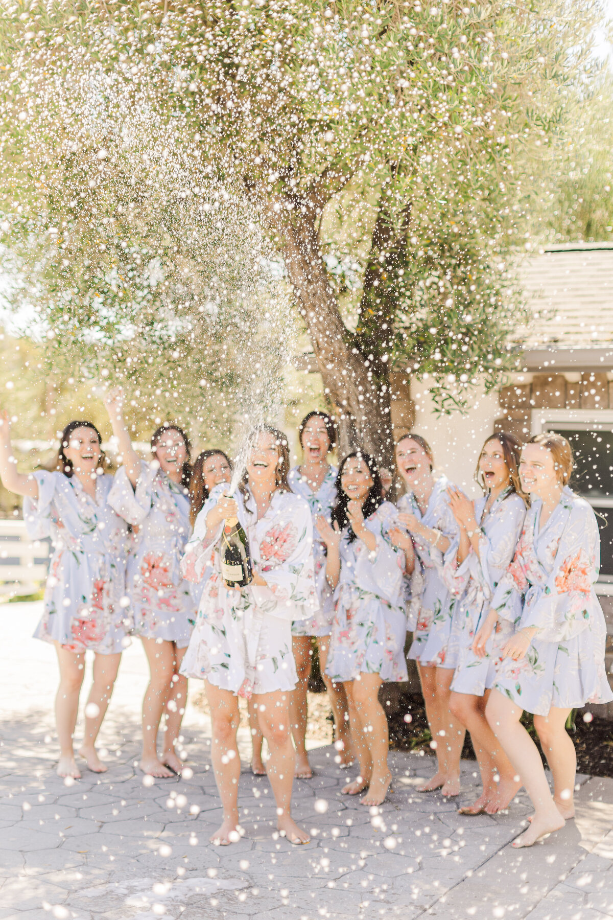 bride and bridesmaids in floral robes popping champagne and laughing together outside.