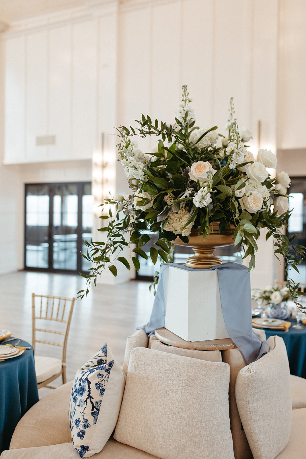 Green and white flower arrangement on gold stand above a white canvased seating area in a banquet room.