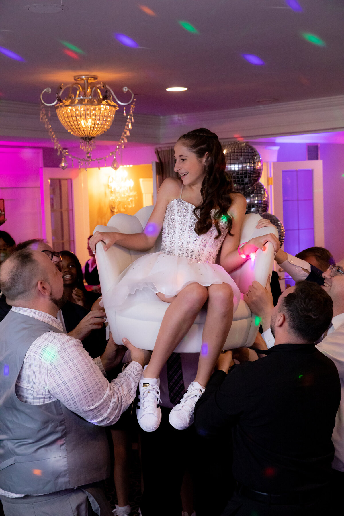 A teenage girl in a white sits in a lifted chair during the horah bat mitzvah Portobello's Oakland New Jersey bar photographer Milestone Images