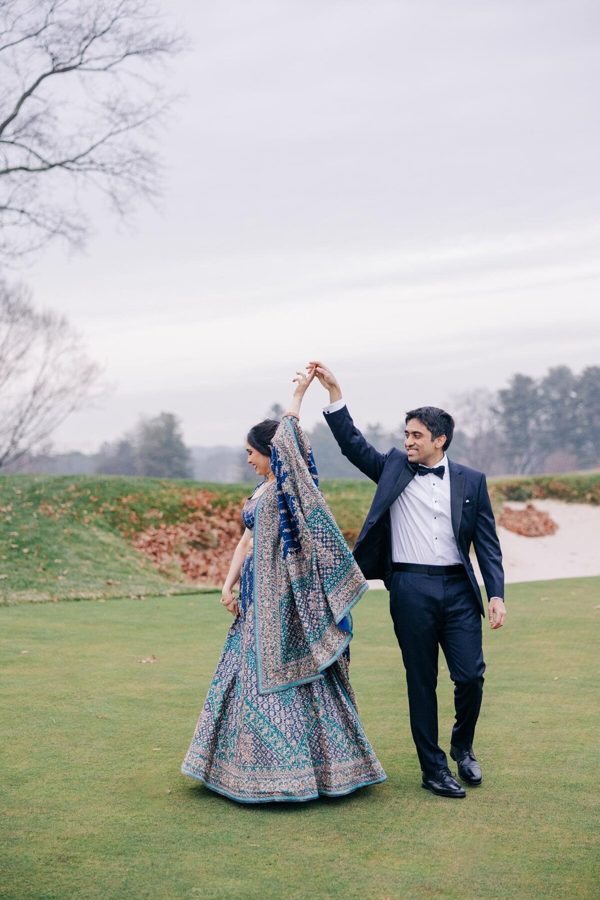 A couple joyfully dances on a golf course; the woman is in a vibrant, embroidered sari, and the man is in a black tuxedo.