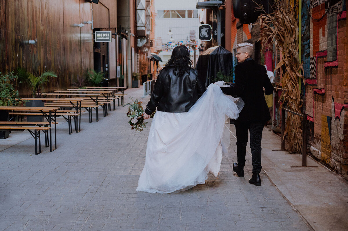 Brides walking down alley while one bride holds her partners dress train for her