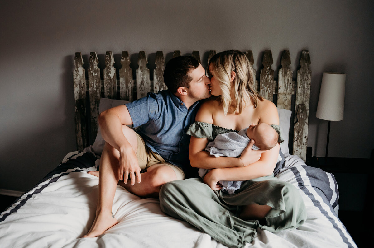 Newborn Photographer, a husband kisses his wife as she holds baby on the bed
