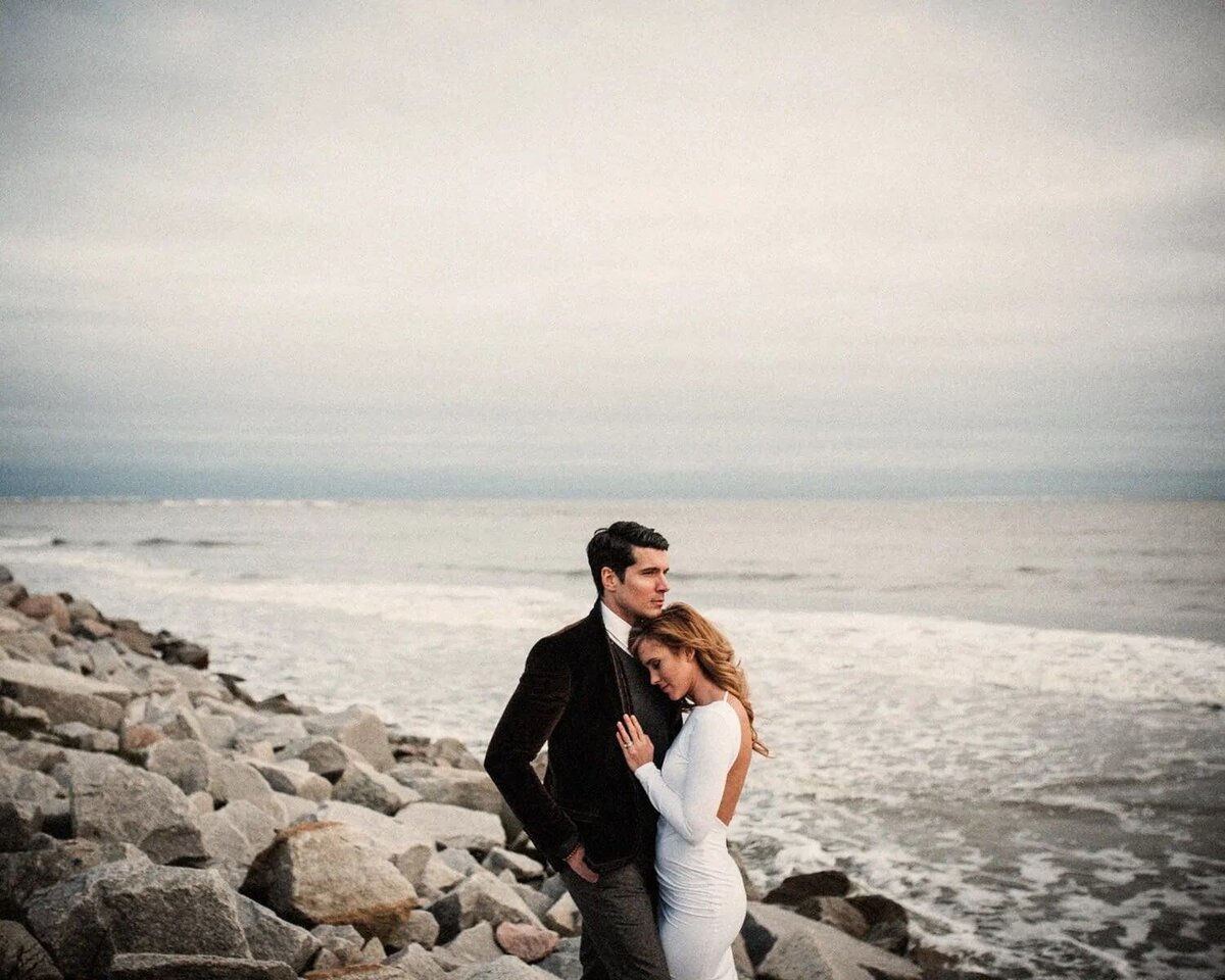 A bride resting her head on a groom's chest while they stand on a rocky coastline.
