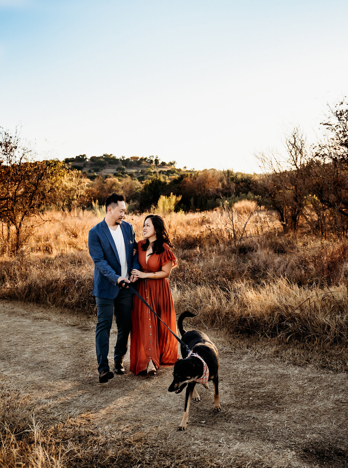 Couples Photography, Man in a blue blazer holding hands with woman in a rust dress as their brown dog walks in front of them, at Commons Ford Ranch Park at sunset.