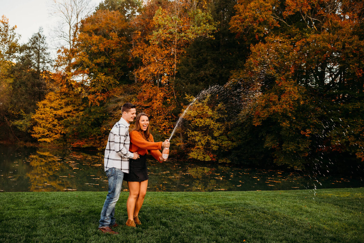 photo of woman popping champagne in front of a pond while her fiance cheers
