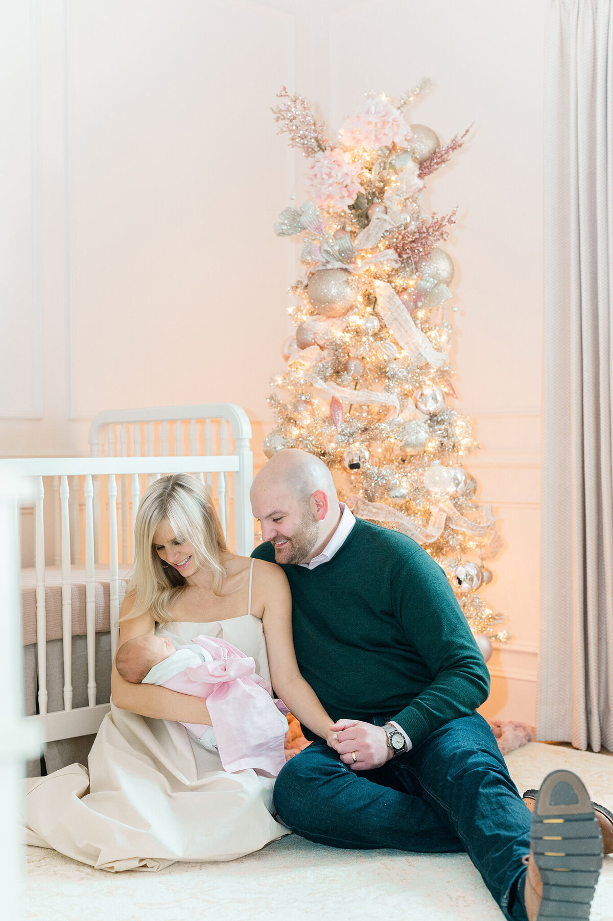 Mother and Father sitting in front of crib with a christmas tree in the background holding a newborn in a Dallas nursery.