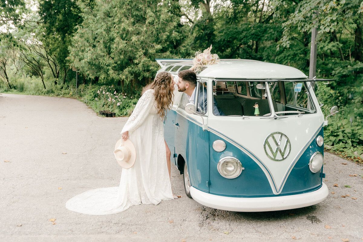 Bride standing by vintage VW bus and the groom is peaking out the the car's window and trying to reach his bride for a kiss