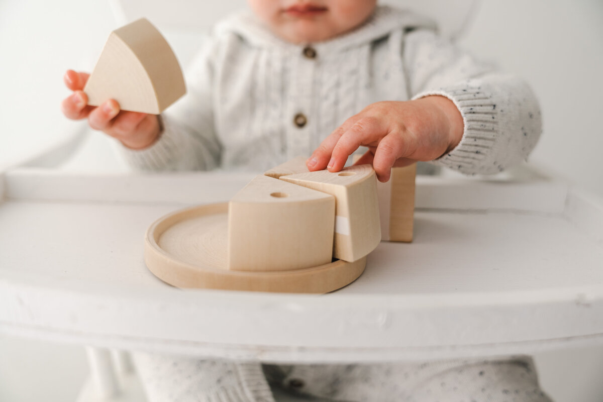 Baby playing with wooden cake on a white highchair. Taken by Fig and Olive Photography, Minneapolis Baby Photographer.