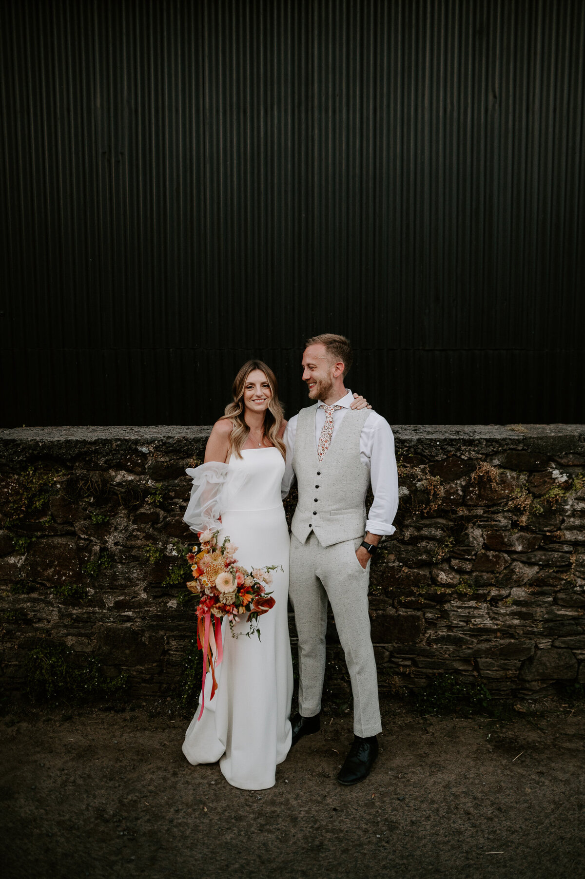 Carrying a large yellow and orange bouquet, a bride stands with her groom outside a black barn at Anran @ Tidwell Farm in Devon.