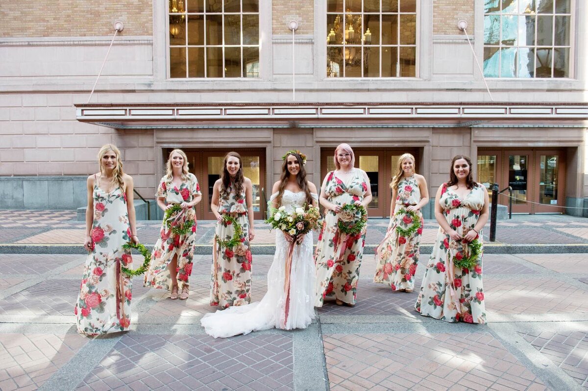 a bride stands in the middle of 6 bridesmaids wearing colorful floral bridesmaid dresses