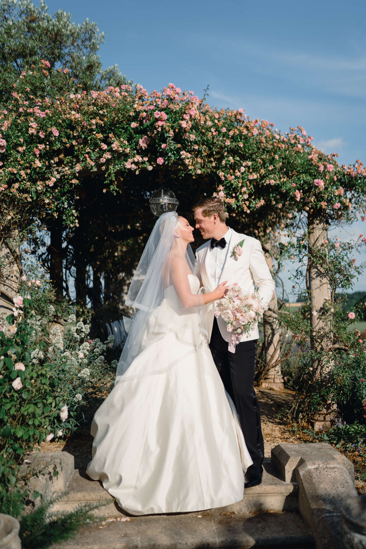 bride and groom embrace in front of a rose arbour covered in pink roses at their romantic cotswold wedding at euridge manor