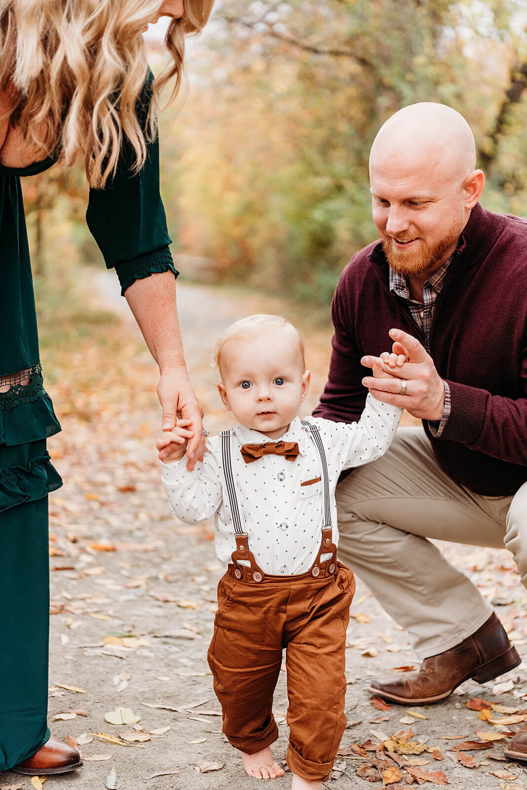 one year old boy walking barefoot wearing rust colored pants, suspenders, and a bowtie on his button down shirt.  He is being supported by holding one hand of each of his parents on each side.