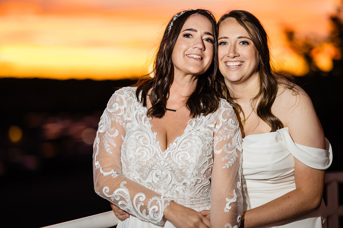 Two brides sharing a tender moment against a sunset, with one bride in a lace gown leaning on the shoulder of the other in an off-the-shoulder dress.