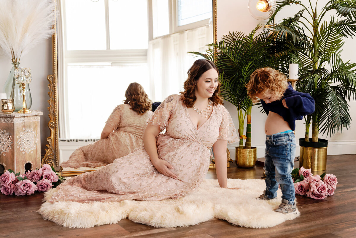 st-louis-maternity-photographer-mom-in-baltic-born-dress-sitting-in-front-of-mirror-with-toddler-boy-lifting-up-his-shirt-to-show-tummy