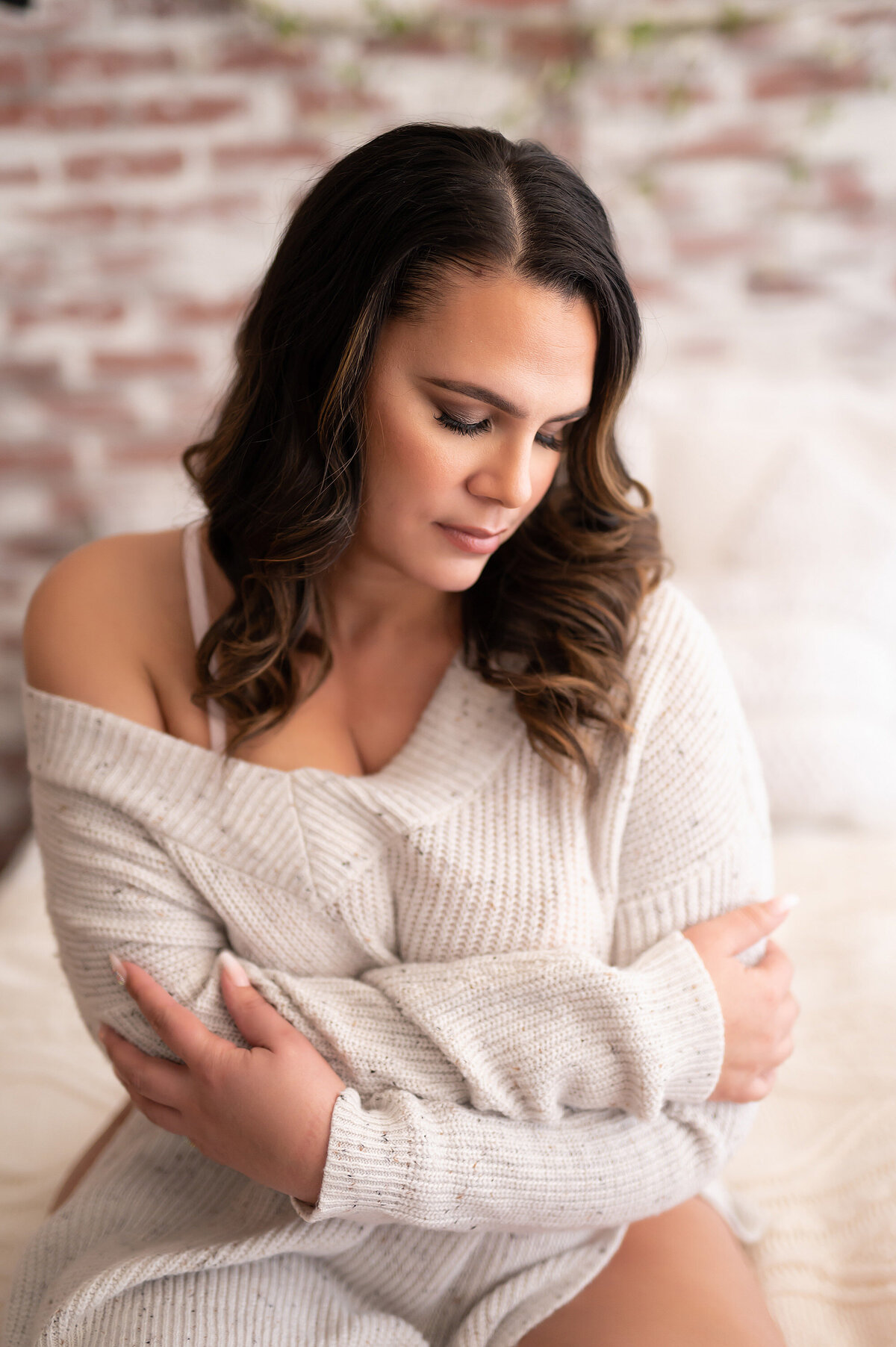 A brunette woman poses in an off-the-shoulder sweater for a cozy  and bright boudoir session.