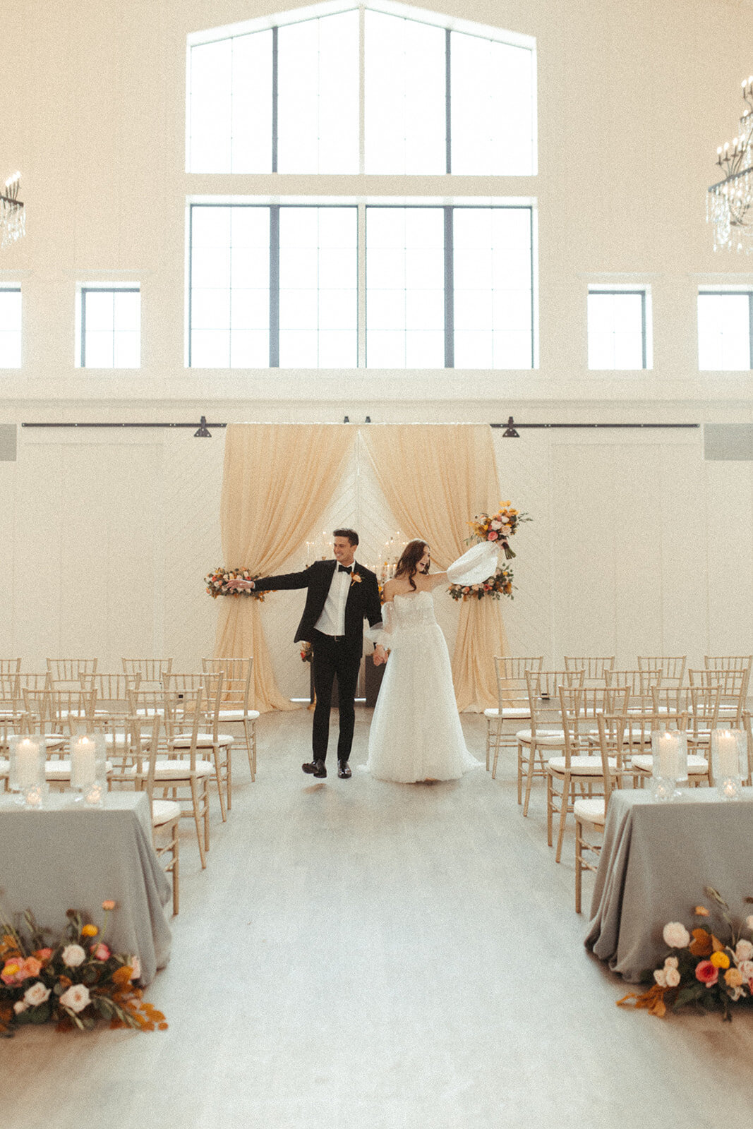 A bride and groom wearing a white wedding gown and black tuxedo walk down the aisle with excitement.