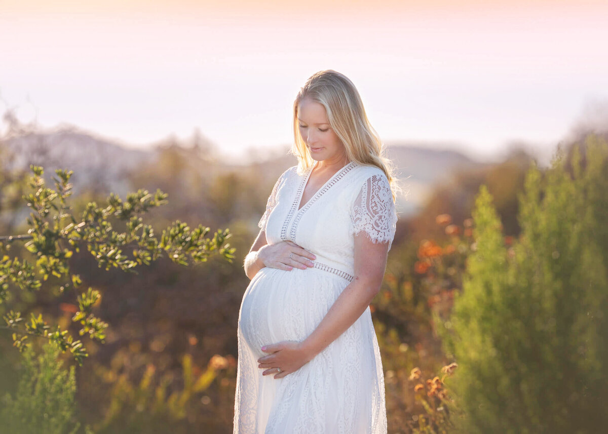 new mom to be photographed in an LA park in a white dress at sunset looking down at baby bump by Los Angeles Maternity Photographer