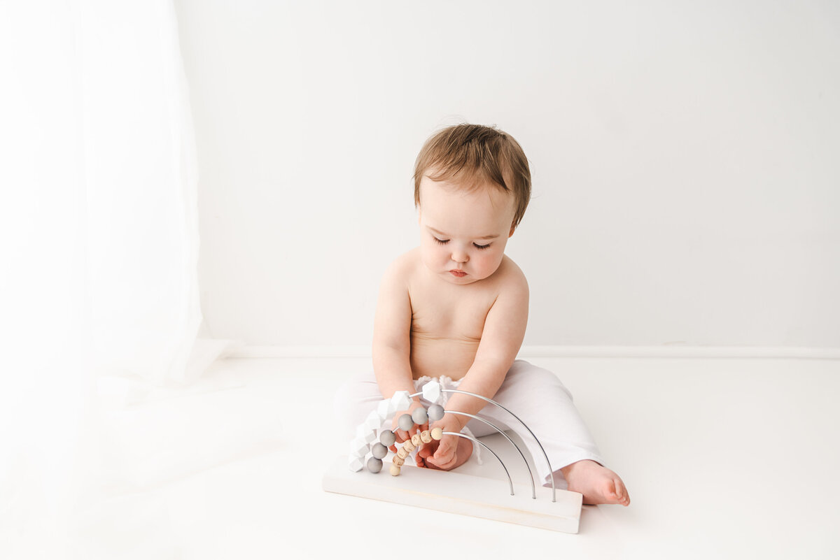 A curious baby sits on a white background, gently touching a strand of wooden beads with a soft focus on discovery and tenderness. Taken by Fig and Olive Photography, Minneapolis Baby Photographer.