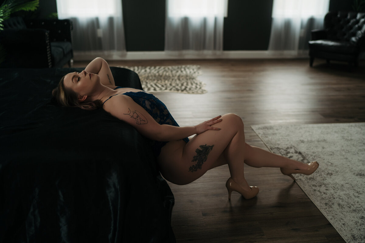 A woman in black lingerie leans back on a clack bed in a studio with hardwood floor