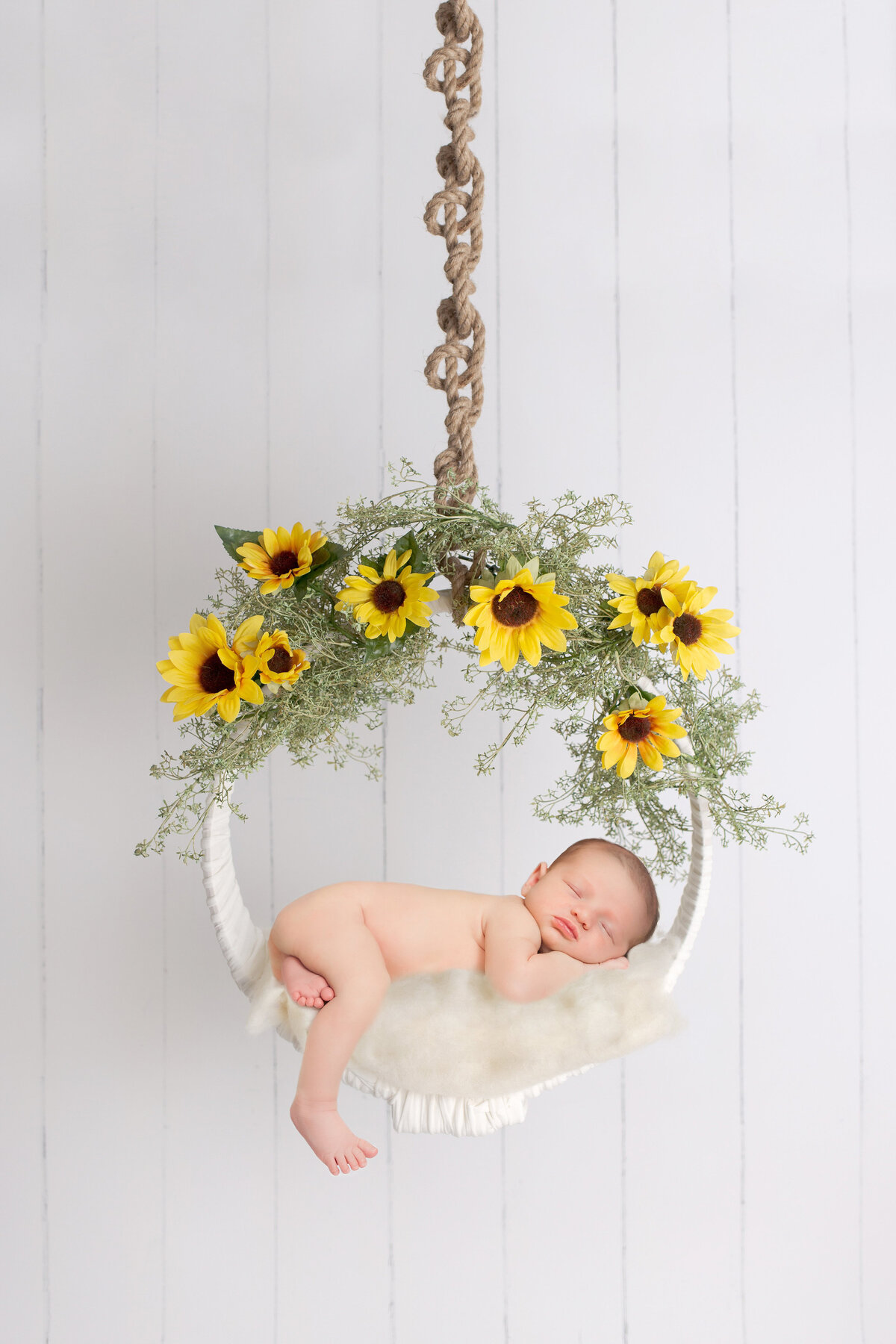 A sleeping baby is curled up on a white, fluffy cushion suspended in a circular swing. The swing, captured by a Jacksonville newborn photographer, is adorned with vibrant sunflowers and greenery, hanging from a thick braided rope against a white, wood-paneled background.