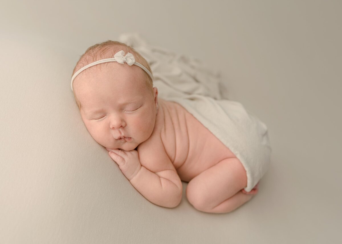 Newborn baby girl in bum up pose laying on beige blanket and matching bow headband and swaddle