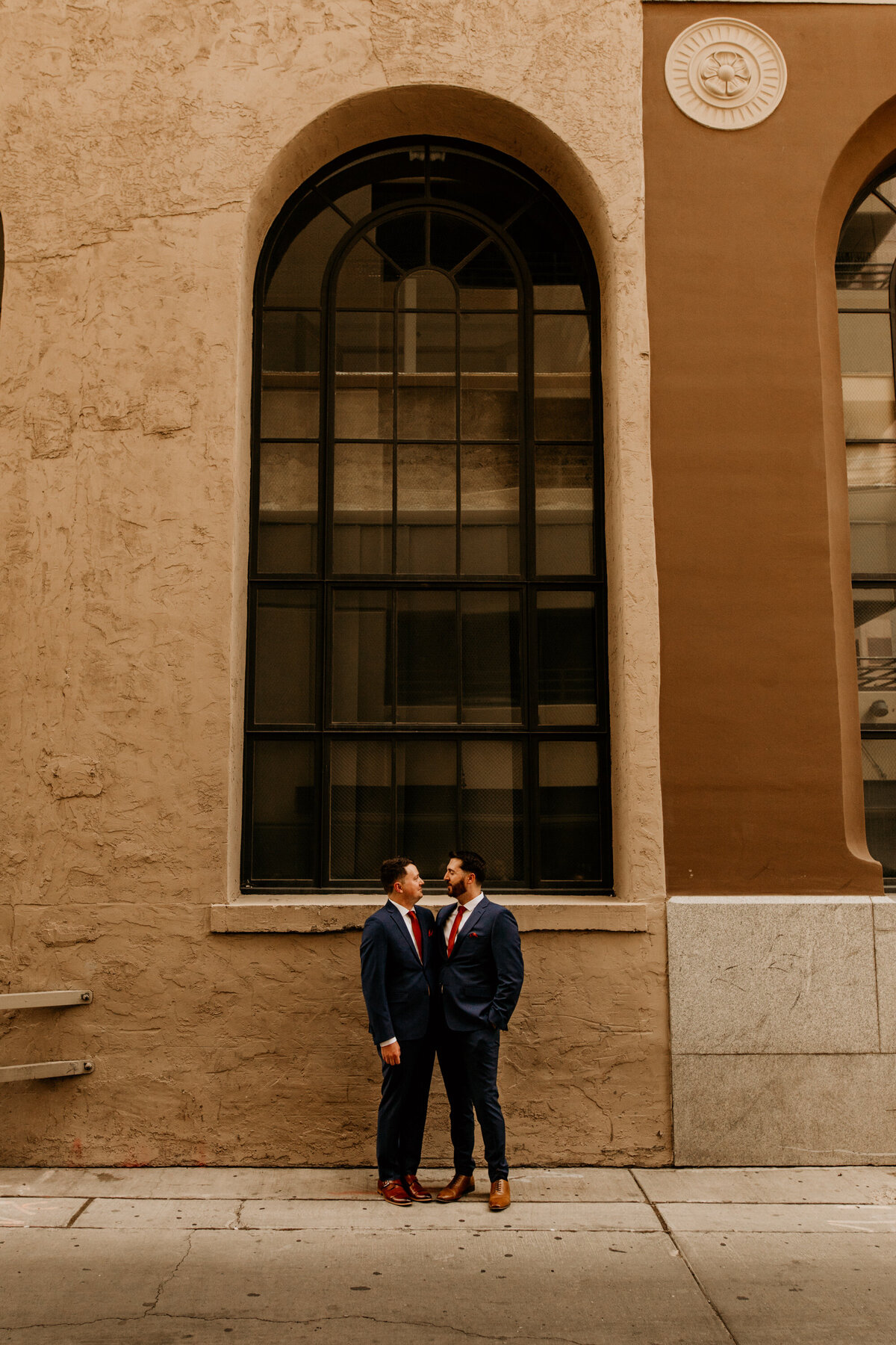 two grooms standing in front of a large window of a brick Albuquerque building