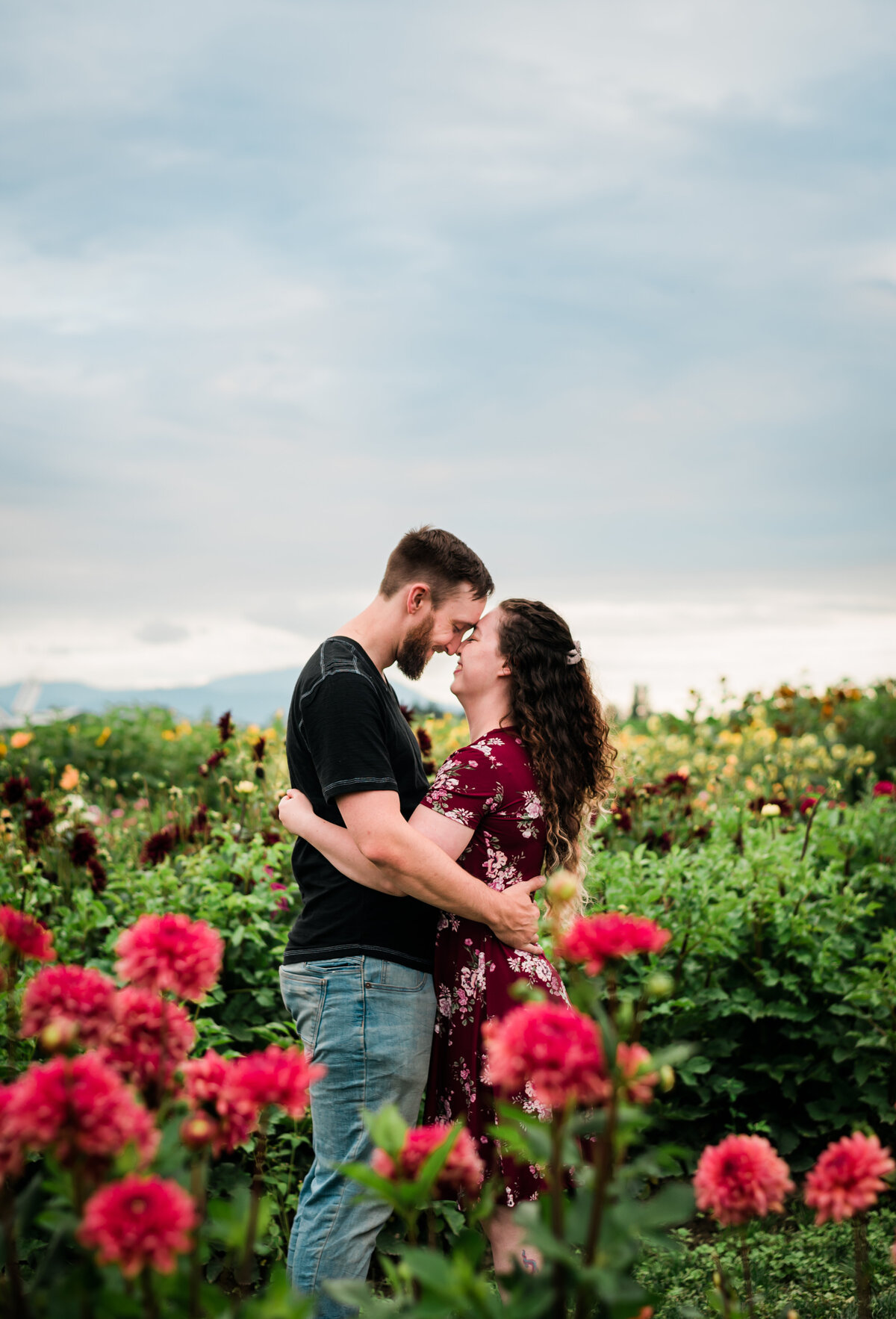 Couple standing in the middle of a flower field. Touching noses together mountain in the back round.