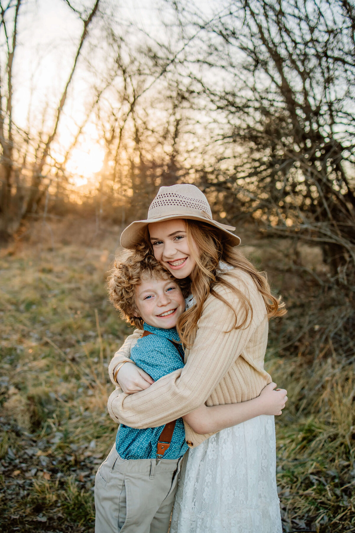 a young boy and girl hugging  closely and  heads are facing the camera big smiles at sunset in a forest