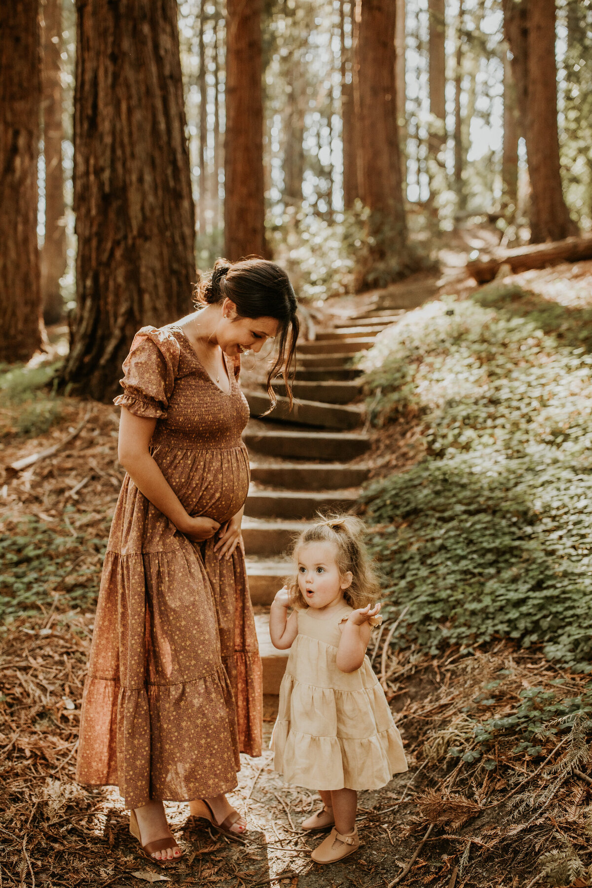 Pregnant mother holds belly and toddler daughter stands at her side making silly face in redwood forest