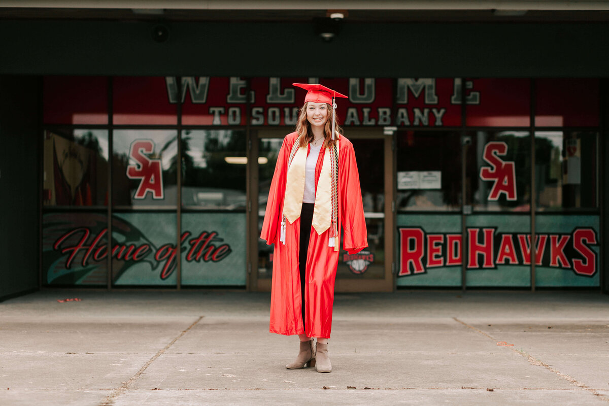 A high school senior in red cap and gown stands in front of South Albany High School