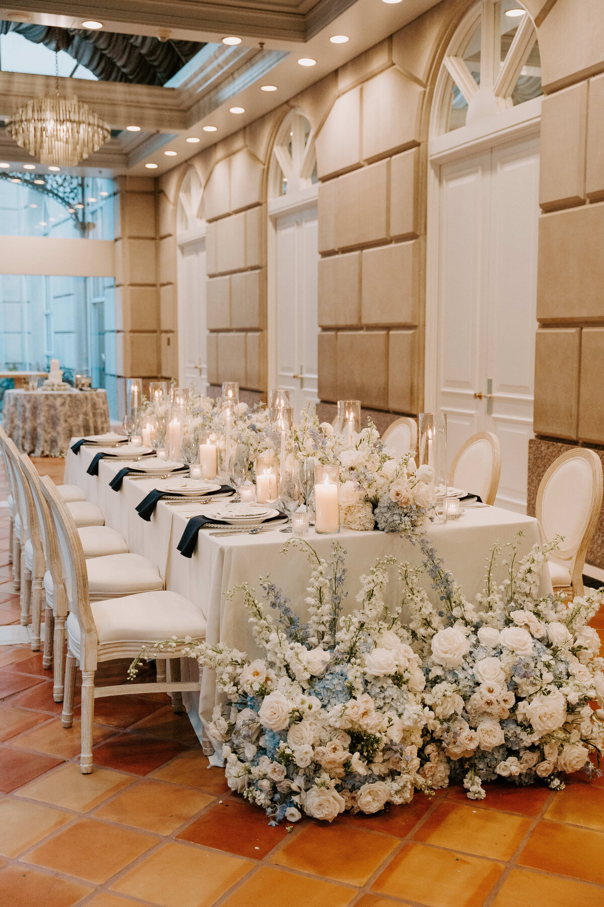 Elegant dining setup with a long table draped in white, adorned with white floral arrangements, candles, and navy napkins. Warm, inviting ambiance in a stylish venue.