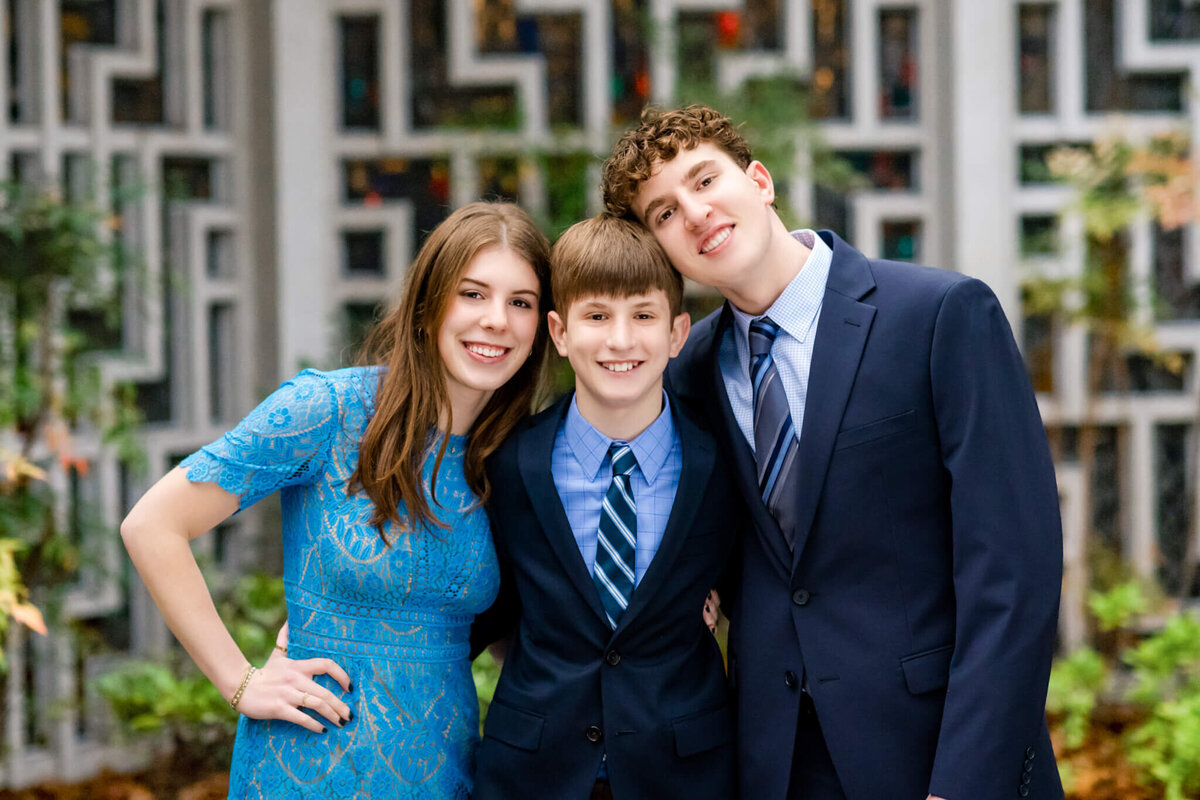 A teenage girl in a blue lace dress stands with her two brothers in blue suits in a garden